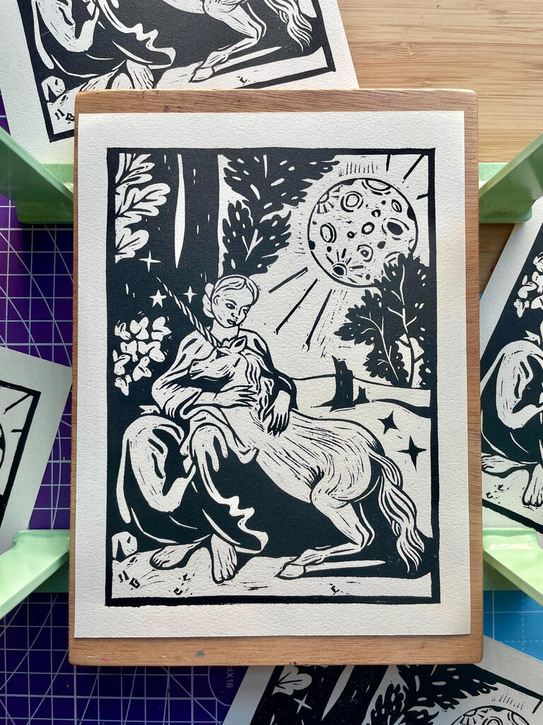 Linocut print of a unicorn resting on a woman's lap, outside under an oversized full moon