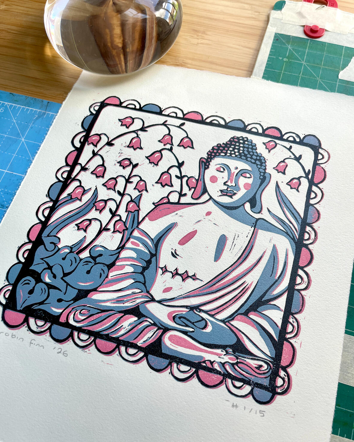 Linocut print of a Buddha statue with top surgery scars, in a garden of Lily of the Valley, surrounded by a decorative border. Printed in pink, blue, and black. Pictured with printmaking tools.