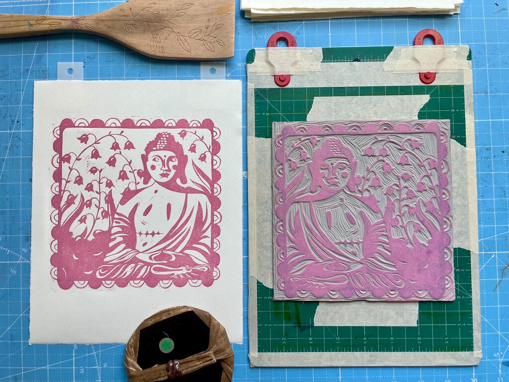A freshly pulled, unfinished linocut print of a Buddha statue with top surgery scars, next to the linoleum block on a cutting mat marked with tape and registration tabs to line up the paper. There are also two tools on the table: a bamboo baren and a woode