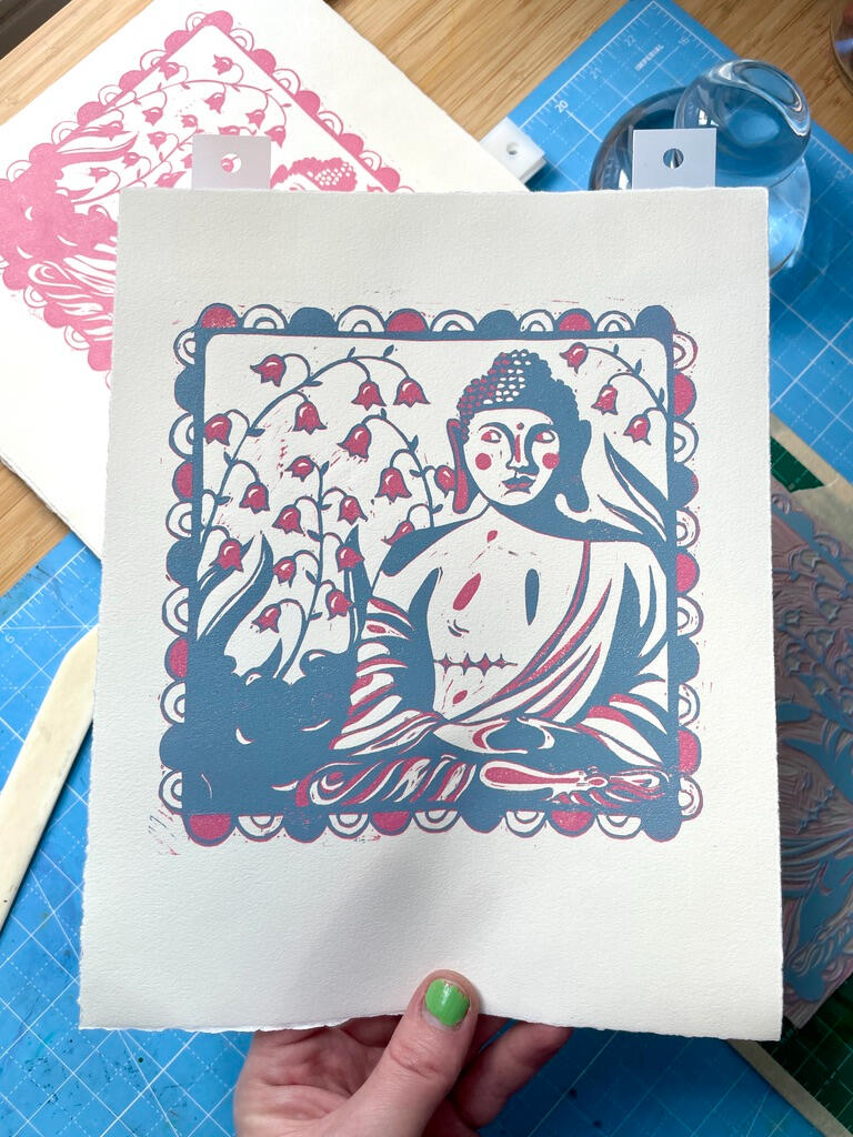 Unfinished, two color linocut block print of a buddha statue with top surgery scars in a garden of lily of the valley. Printmaking tools in the background.