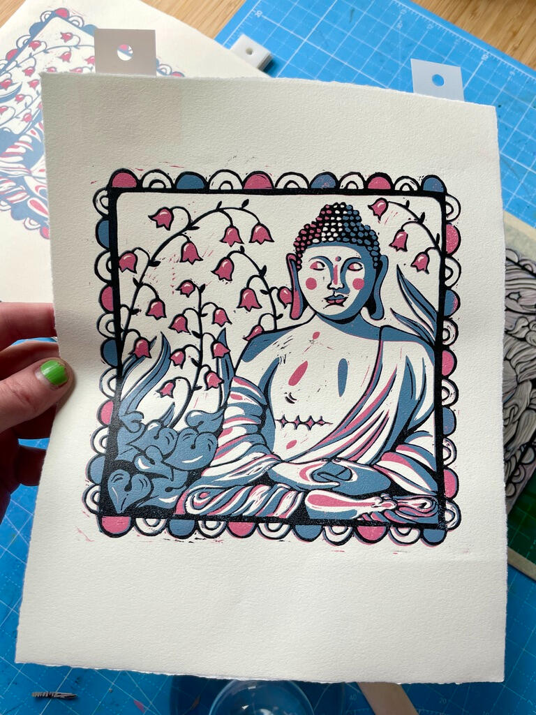 A linocut print of a Buddha statue with top surgery scars in a garden of lily of the valley, with a decorative border. Printmaking supplies in the background.