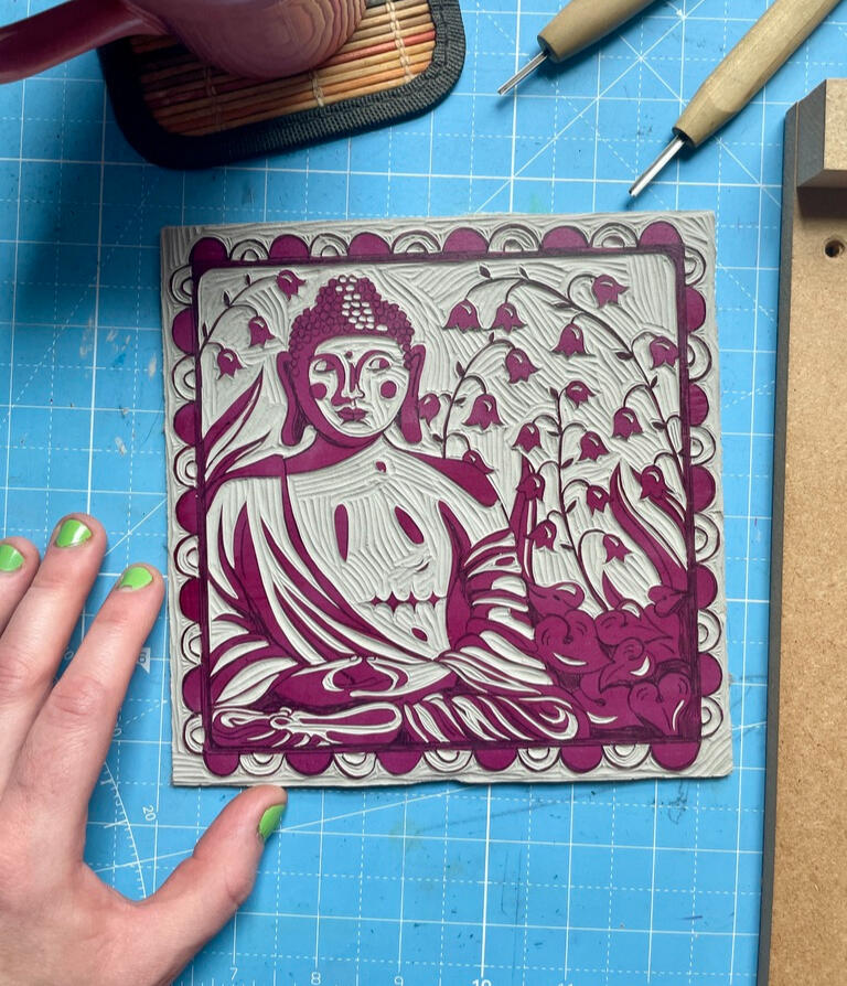 A carved linoleum block on a cutting mat next to carving tools, a mug of tea, and Robin's hand. The design is a buddha statue with top surgery scars surrounded by lily of the valley flowers, leaves, and a decorative border.